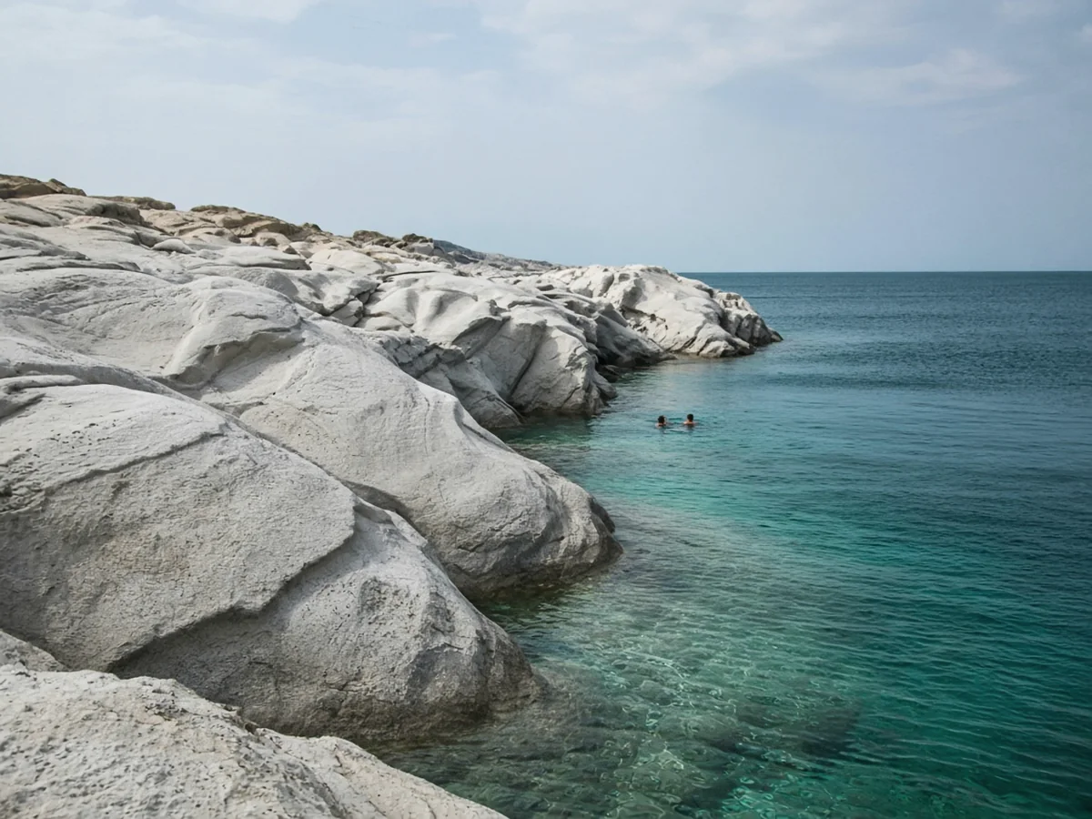 Best Greek Islands for First-Time Visitors Smooth pale white-grey rock formations curving down to clear turquoise water, with two small swimmers in the inlet