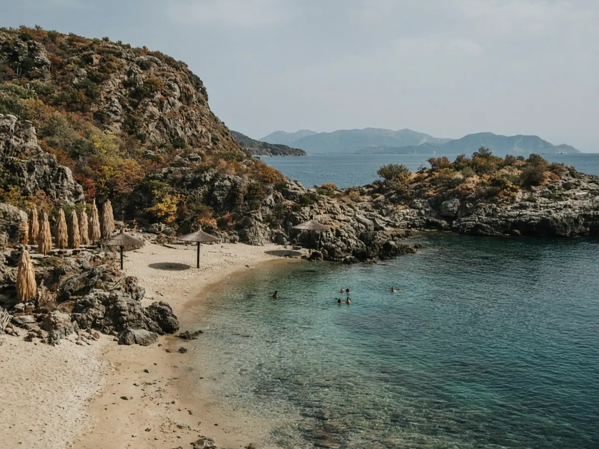 A sheltered sandy cove under a soft autumn sky, with a few swimmers in calm clear water, straw umbrellas mostly folded, and rocky headlands with autumn-tinted scrub
