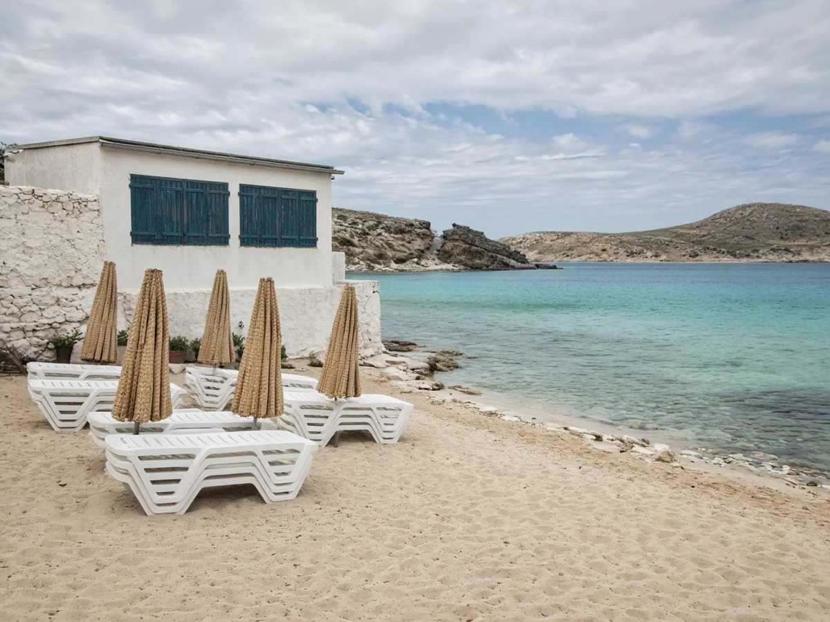 An empty beach at the end of the season: white sunbeds stacked in piles, folded straw umbrellas and a small white beach bar with its dark teal shutters closed beside a calm turquoise sea