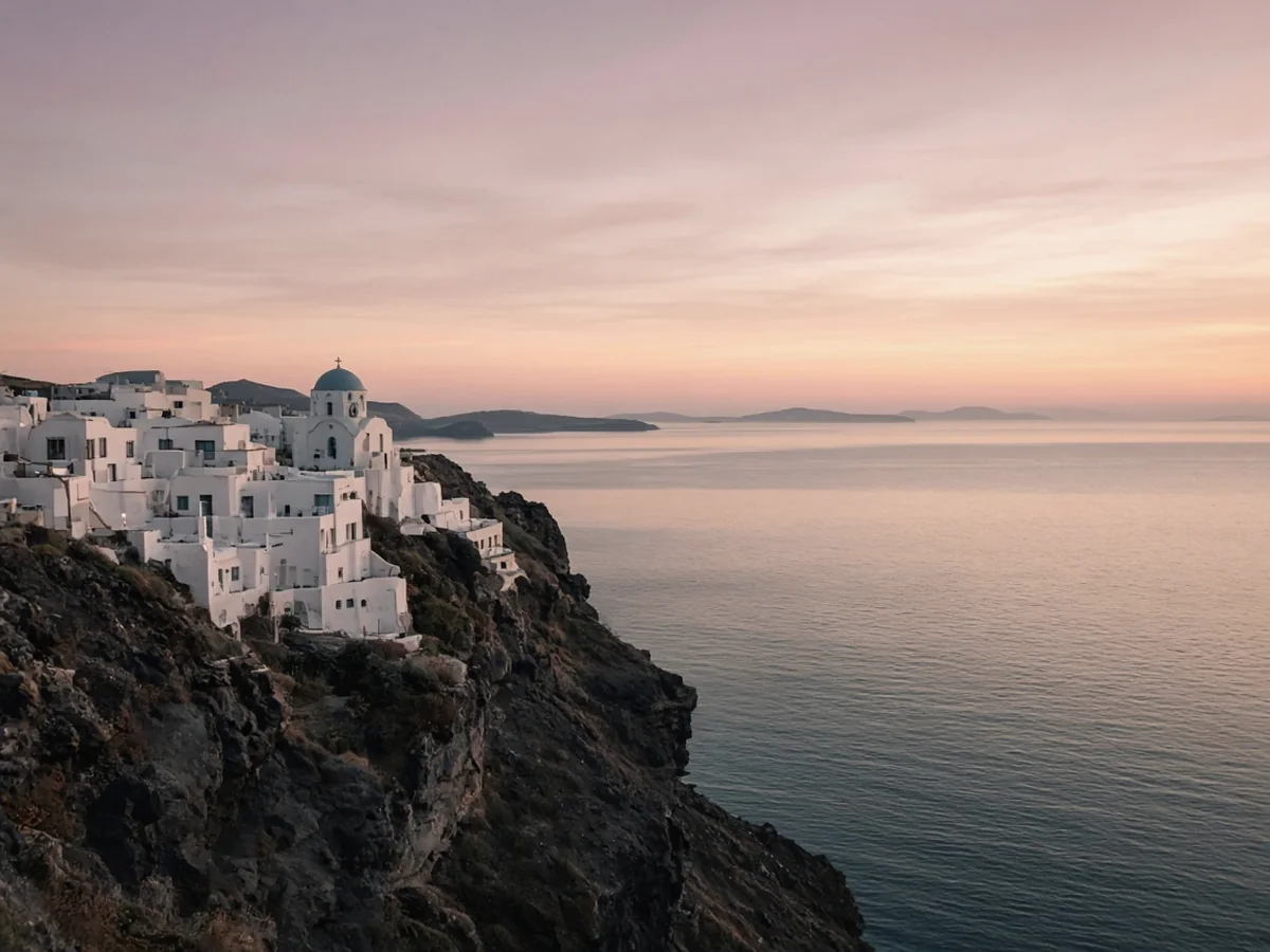 Whitewashed houses and a small chapel with a blue-grey dome on the edge of a dark cliff above a calm bay at sunset, under a pink and apricot sky