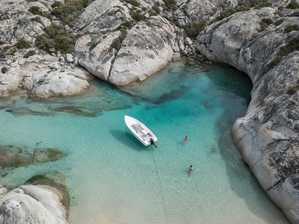 Aerial view of a small white motor boat anchored in a clear turquoise cove between smooth pale rocks, with two swimmers nearby