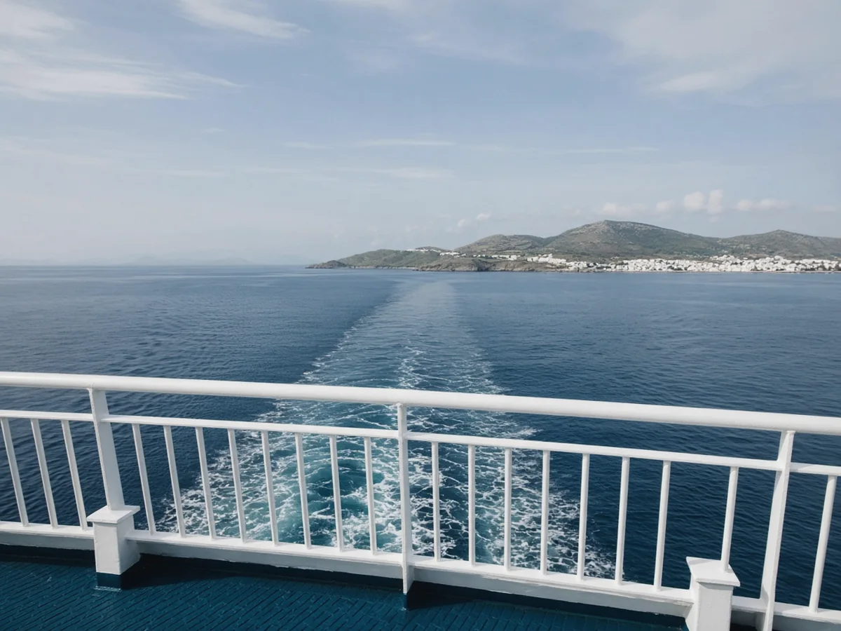 Naxos vs Paros vs Milos: Which Cyclades Island Fits Your Trip? The view from a ferry's upper deck: a white railing, a long wake across deep blue sea and a hilly island with a white town on the horizon