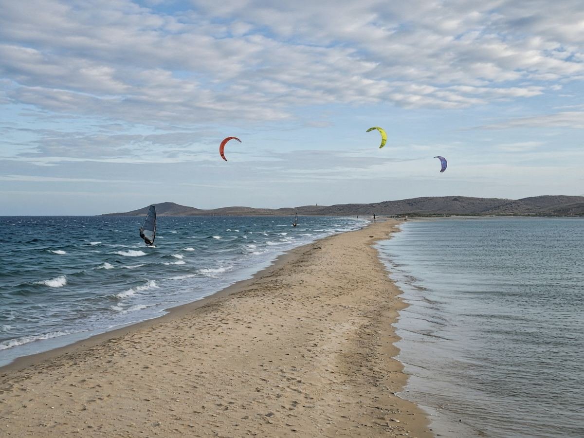 Rhodes Travel Guide A long narrow sandy spit between choppy water and calmer water, with a windsurfer and three kitesurfing kites in the air under a cloud-streaked sky