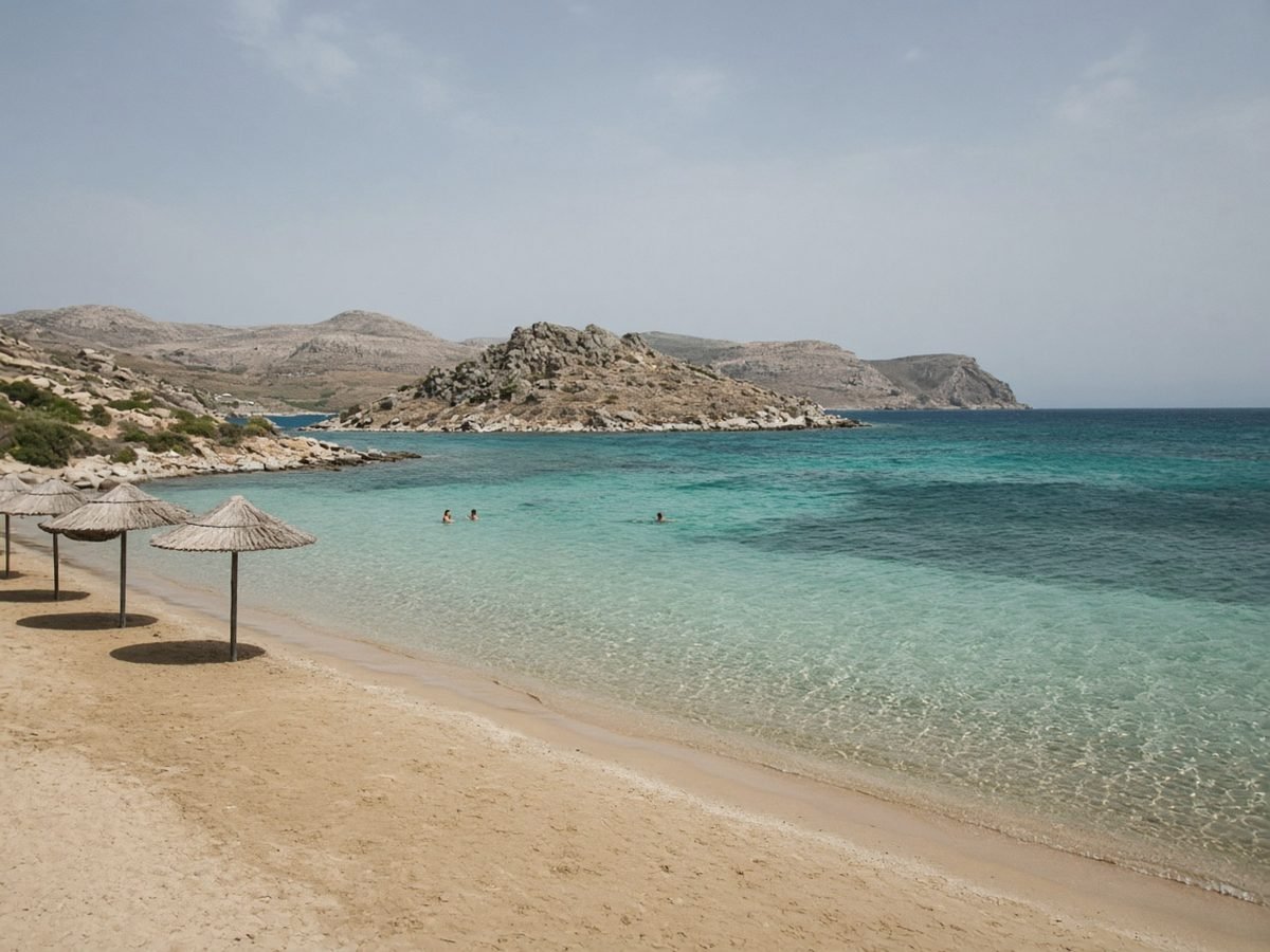 Syros Travel Guide A quiet sandy beach with straw umbrellas and clear, shallow turquoise water, with a few swimmers and a small rocky islet just offshore