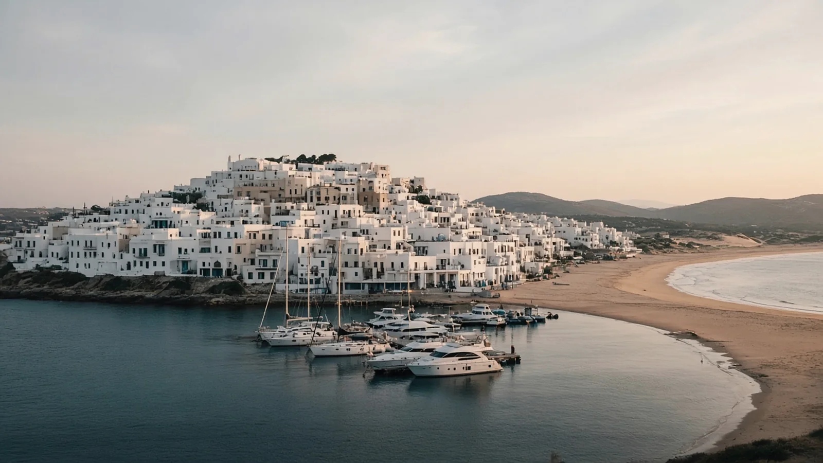 Where to Stay in Naxos A whitewashed town climbing a low hill above a small marina with yachts in soft evening light, with a long sandy beach curving away to one side
