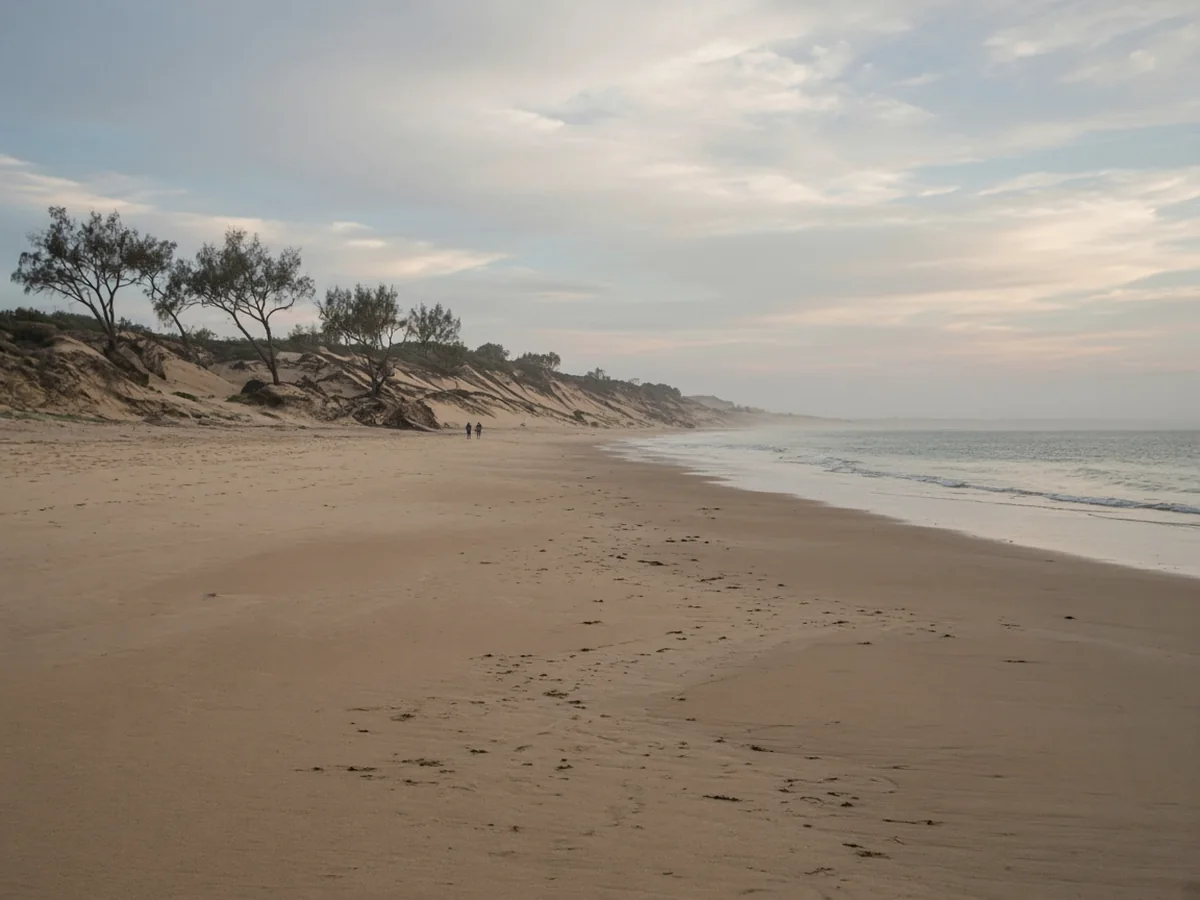 Where to Stay in Naxos A wide, almost empty sandy beach in soft evening light, with low dunes and scattered trees behind and two people walking far down the shore