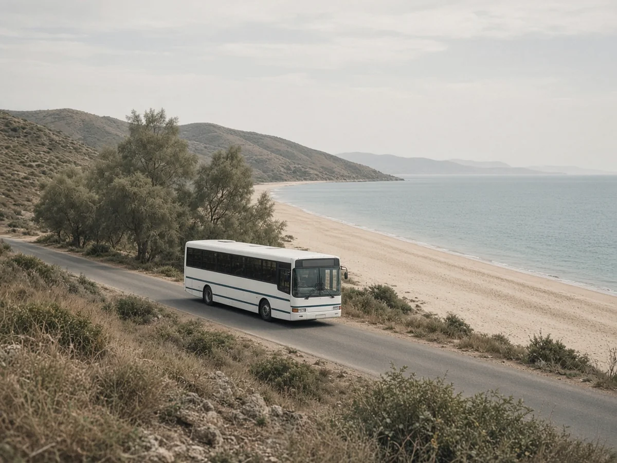 Where to Stay in Naxos A plain white bus on a coastal road beside a long sandy beach, with trees, dry hills and a calm sea