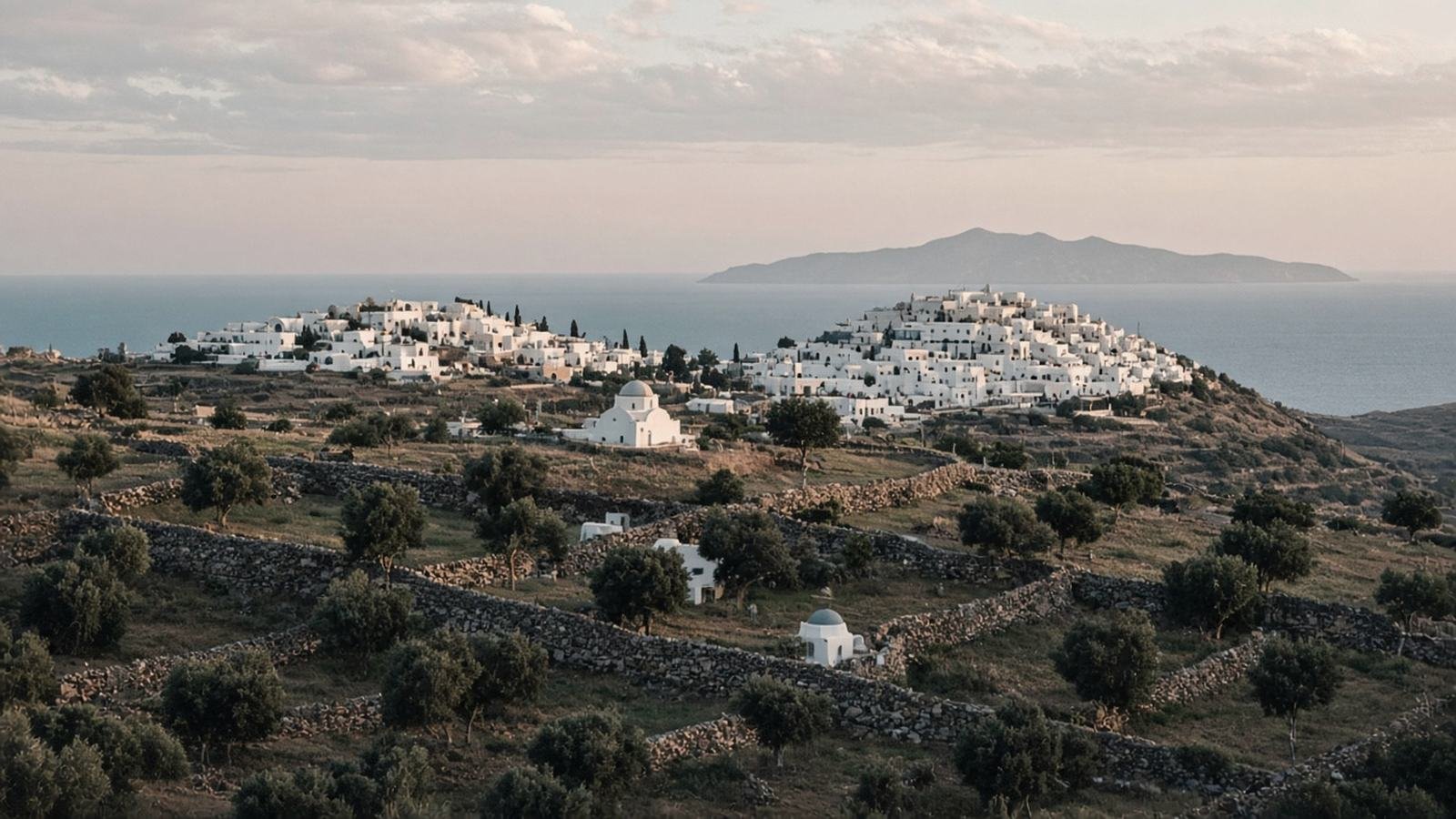 Two white villages spread across a gentle hilltop in early-evening light, above terraced fields with dry-stone walls, olive trees and small chapels, with the sea and a distant island beyond