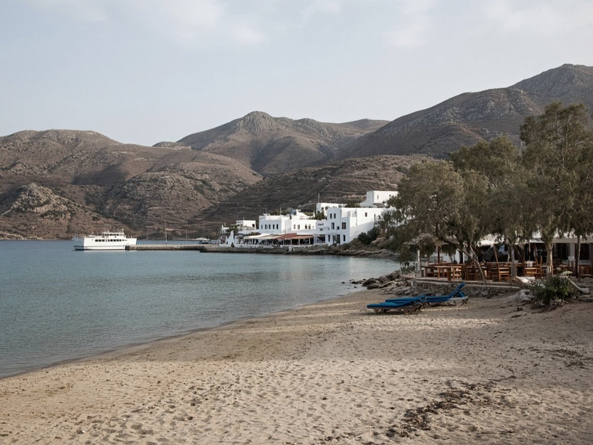 A sandy beach at the head of a deep bay, with tamarisk trees, waterfront tavernas and sunbeds, a small white ferry at the quay and bare brown mountains around