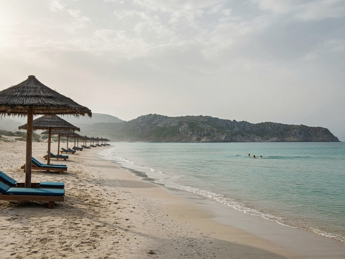 A long sandy beach with straw umbrellas and sunbeds along calm, shallow turquoise water, with a few swimmers and a low rocky headland across the bay
